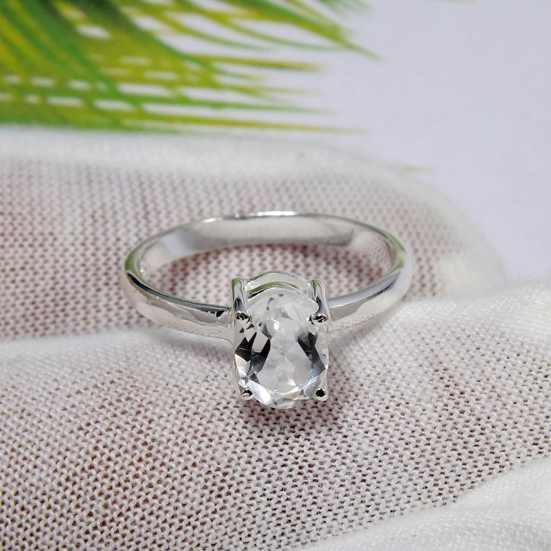 May include: A silver ring with a clear oval-shaped gemstone. The ring is on a white fabric background.