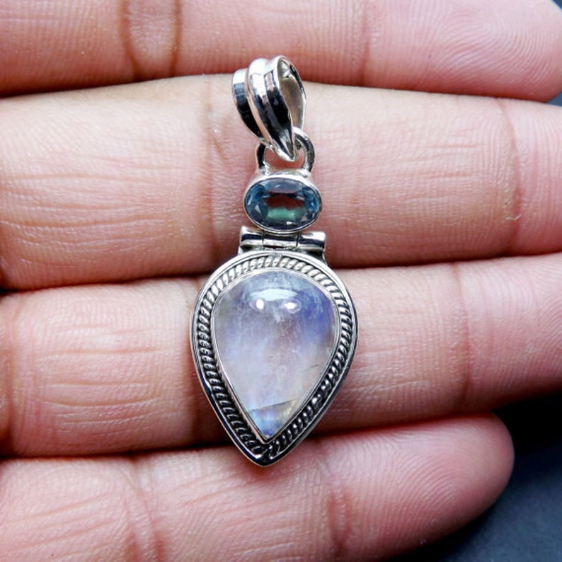 May include: A silver pendant with a teardrop-shaped moonstone and a smaller blue topaz gemstone. The pendant has a detailed silver design around the stones.