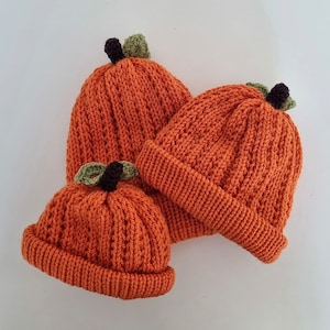May include: Three knitted pumpkin hats in orange with green stems and brown tops. The hats are different sizes.