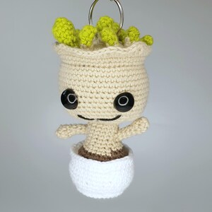 May include: A crocheted plush toy of a character with a green leafy head, brown eyes, and a white pot. The toy is made of yarn and has a smiling expression.