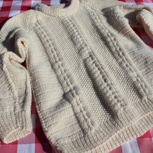 May include: A cream-coloured knitted jumper with a cable knit pattern. The jumper has long sleeves and a crew neck.