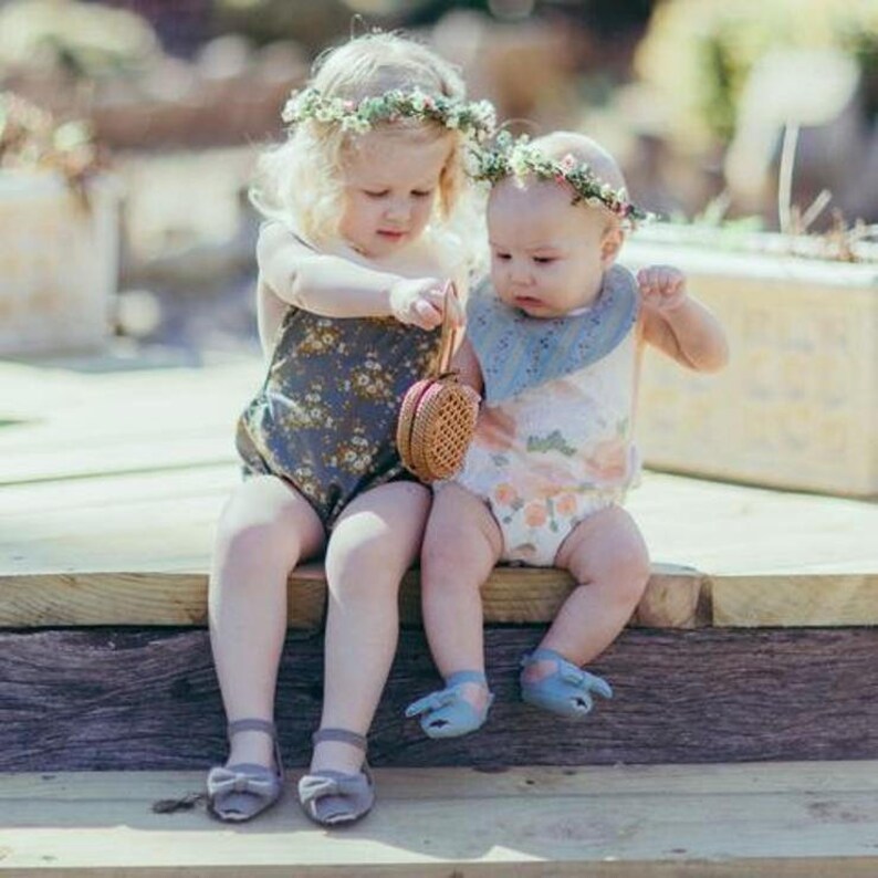 May include: Two young children wearing floral crowns and rompers sit on a wooden surface. One child holds a small, woven purse. The children wear matching gray shoes with bows. The background is blurred, suggesting an outdoor setting.
