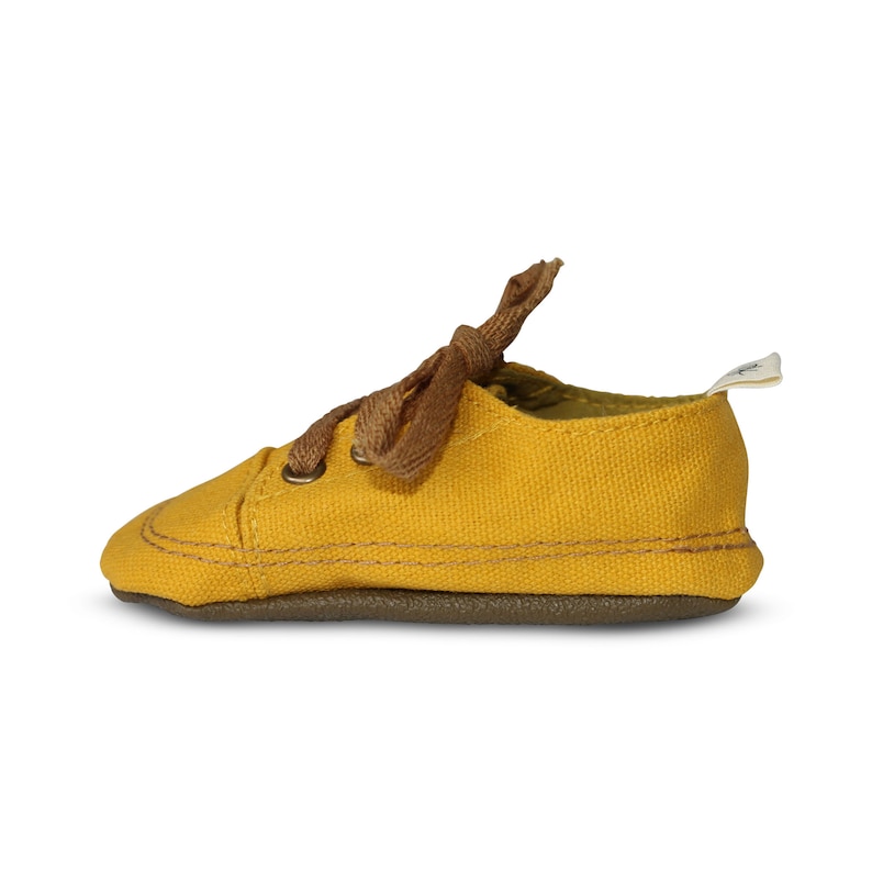 May include: A pair of yellow canvas baby shoes with brown laces. The shoes have a brown rubber sole.