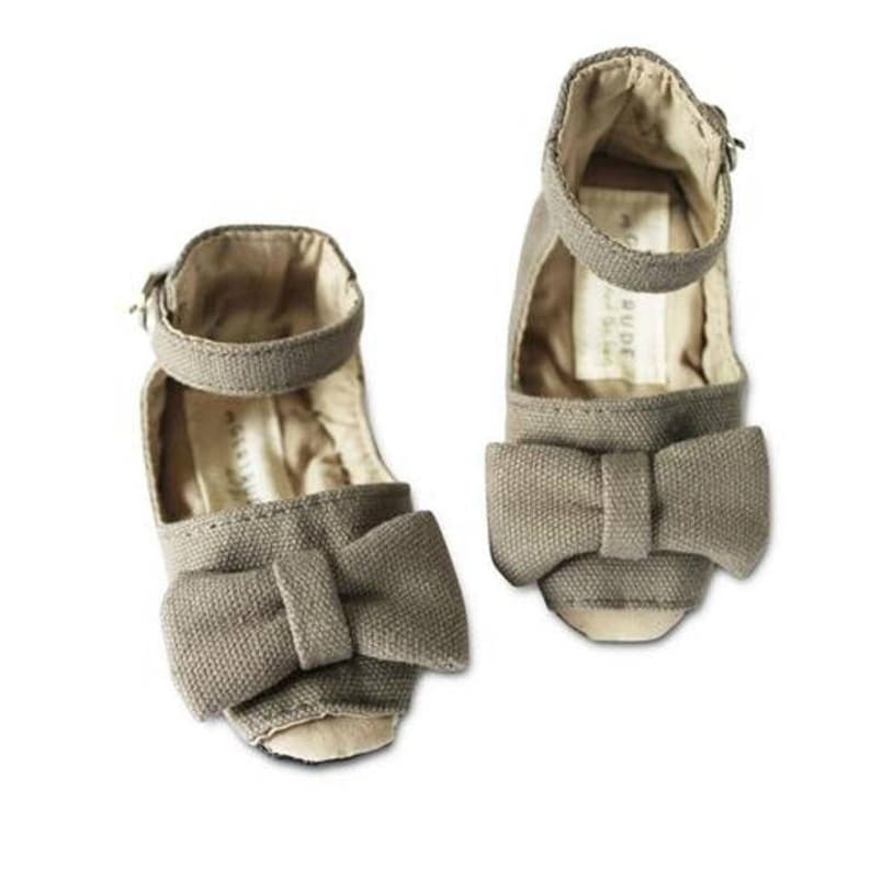 May include: Two pairs of taupe-colored canvas baby shoes. Each shoe features a large bow on the toe and an ankle strap with a buckle. The interior of the shoes is a light beige color.