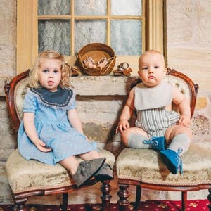 May include: Two children sit on antique chairs. One wears a light blue dress with a dark blue collar and gray shoes. The other wears a striped romper, a bib, gray socks, and blue shoes. A woven basket and shoes sit on a ledge.