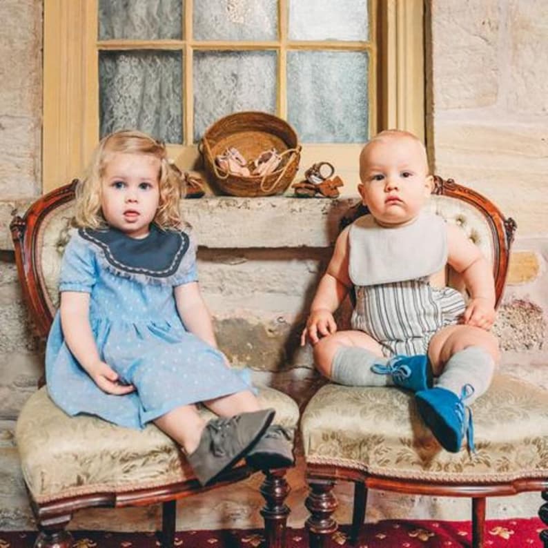 May include: Two children sit on antique chairs. One wears a light blue dress with a dark blue collar and gray shoes. The other wears a striped romper, a bib, gray socks, and blue shoes. A woven basket and shoes sit on a ledge.