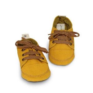 May include: A pair of yellow canvas baby shoes with brown laces. The shoes have a simple design and are perfect for a baby's first steps.
