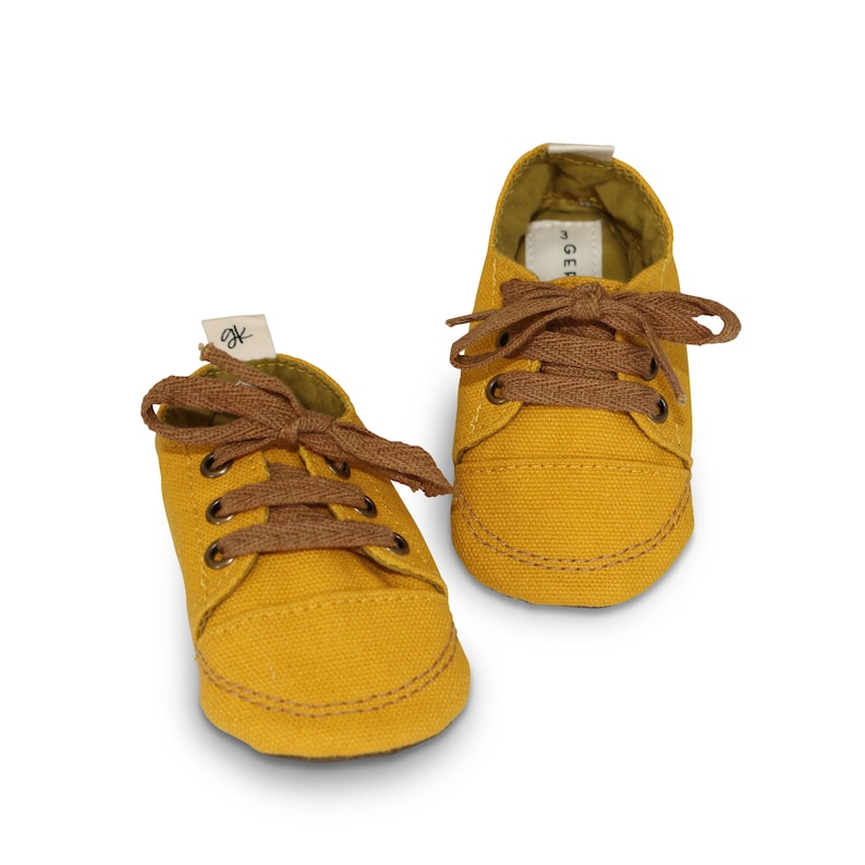 May include: A pair of yellow canvas baby shoes with brown laces. The shoes have a simple design and are perfect for a baby's first steps.