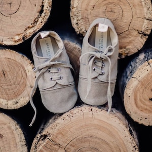 May include: A pair of light brown canvas baby shoes with laces, resting on a stack of wooden logs. The shoes have the text "Gertrude" printed on the inner sole. The shoes are designed with a classic sneaker style.