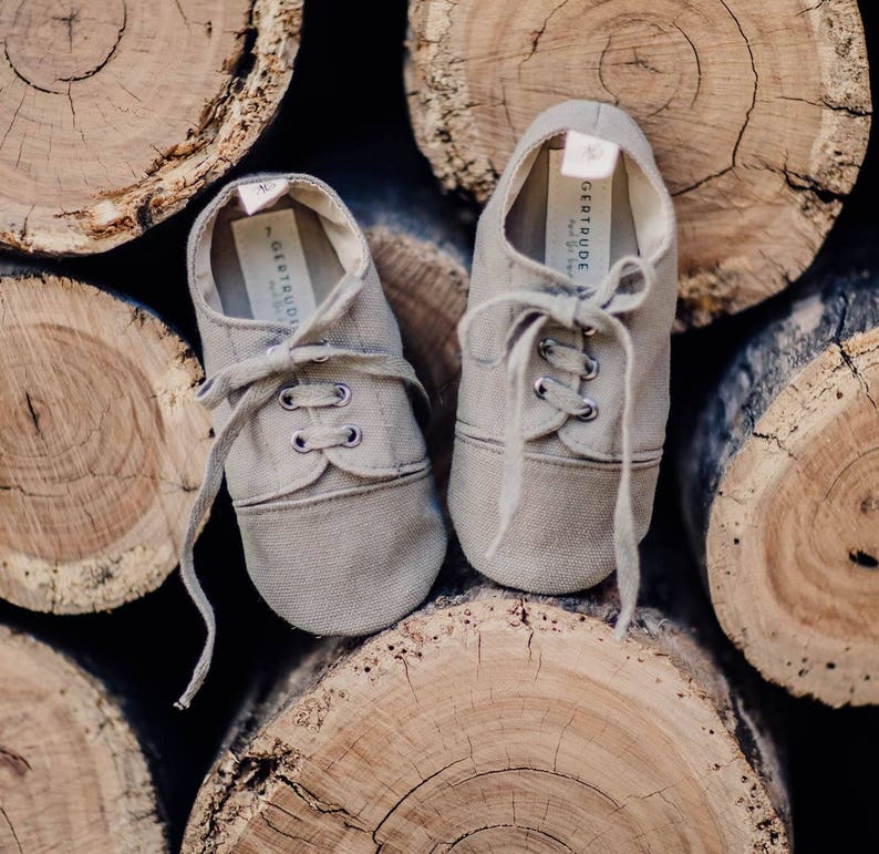 May include: A pair of light brown canvas baby shoes with laces, resting on a stack of wooden logs. The shoes have the text "Gertrude" printed on the inner sole. The shoes are designed with a classic sneaker style.