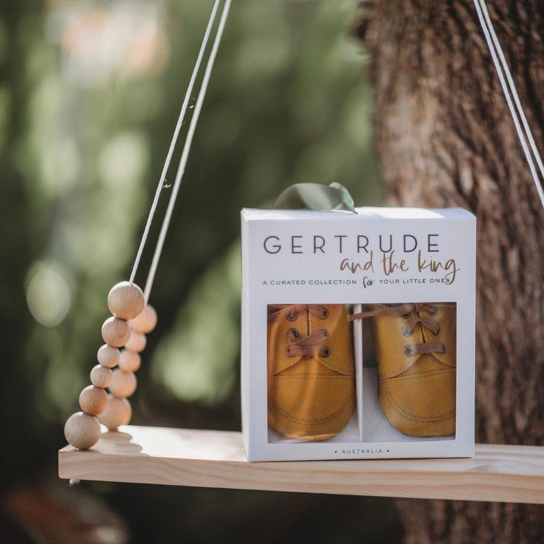 May include: A white box with the text "GERTRUDE and the king" and "A CURATED COLLECTION for YOUR LITTLE ONES" on it. The box contains a pair of yellow leather baby shoes with laces.