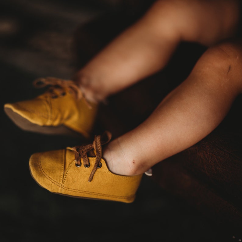 May include: A pair of yellow canvas shoes with brown laces. The shoes are worn by a child and are sitting on a brown surface.
