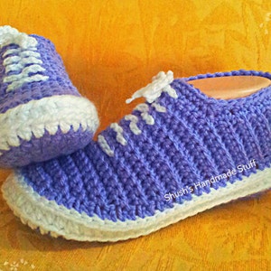 May include: A pair of purple crocheted slippers with white trim. The slippers have a lace-up design and are made with a soft, comfortable yarn.