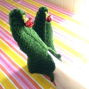 May include: A pair of green crocheted slipper socks with red jingle bells attached. The socks are on a striped pink and yellow surface.