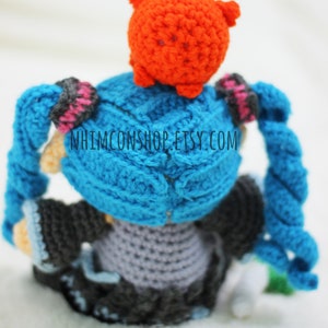 May include: A crocheted anime character with blue hair and a red hat. The character is wearing a gray dress and black shoes. The character is sitting on a white surface. The text "NHIMCONSHOP.ETSY.COM" is visible on the character's back.