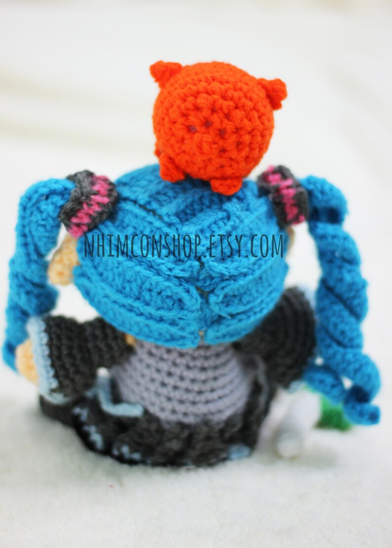 May include: A crocheted anime character with blue hair and a red hat. The character is wearing a gray dress and black shoes. The character is sitting on a white surface. The text "NHIMCONSHOP.ETSY.COM" is visible on the character's back.
