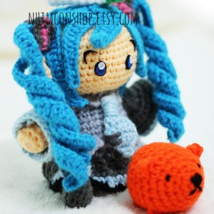 May include: A crocheted anime character with blue hair and a light blue and gray outfit. The character is holding a small orange crocheted animal.