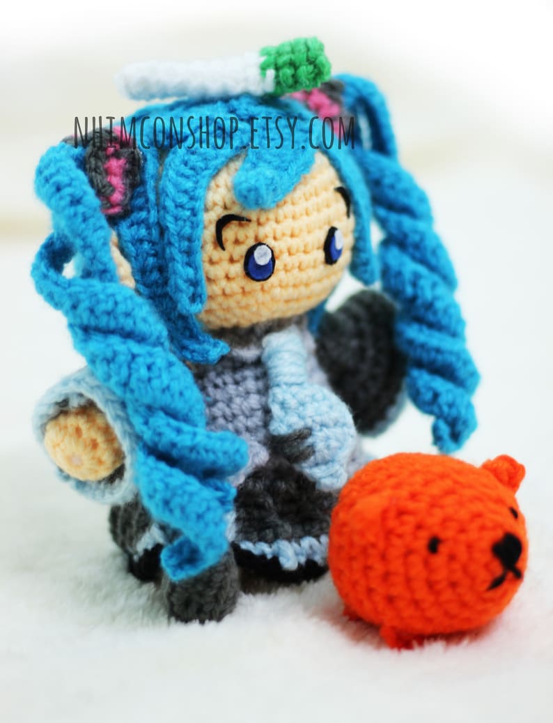 May include: A crocheted anime character with blue hair and a light blue and gray outfit. The character is holding a small orange crocheted animal.