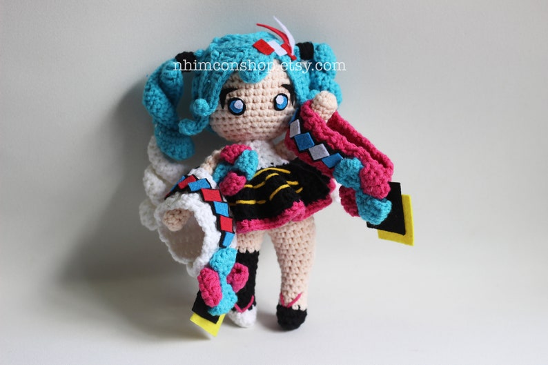 May include: Crocheted plush toy of a female anime character with blue hair, wearing a white, pink, and black outfit with a blue and pink scarf. The toy is holding a yellow and black object in its right hand. The toy is standing on a white surface. The text "nhimconshop.etsy.com" is visible in the background.
