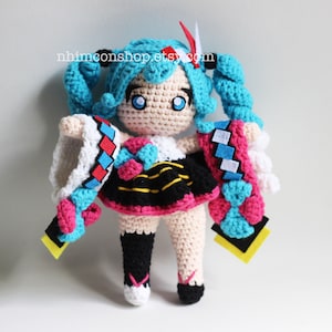 May include: Crocheted plush toy of a female anime character with blue hair, a white and red dress, and a yellow and black skirt. The toy is holding a blue and pink crocheted scarf in each hand. The toy is standing on a white surface. The text "nhmconshop.etsy.com" is visible in the background.
