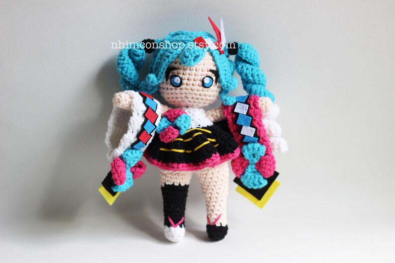 May include: Crocheted plush toy of a female anime character with blue hair, a white and red dress, and a yellow and black skirt. The toy is holding a blue and pink crocheted scarf in each hand. The toy is standing on a white surface. The text "nhmconshop.etsy.com" is visible in the background.
