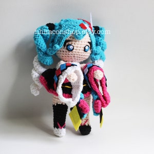 May include: Crocheted plush toy of a female anime character with blue hair, wearing a white, black, and pink outfit. The toy is holding a pink and white crocheted accessory. The toy is standing on a white surface. The text "nhinconshop.etsy.com" is visible in the background.
