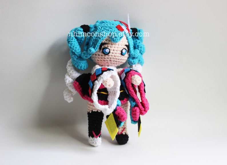 May include: Crocheted plush toy of a female anime character with blue hair, wearing a white, black, and pink outfit. The toy is holding a pink and white crocheted accessory. The toy is standing on a white surface. The text "nhinconshop.etsy.com" is visible in the background.