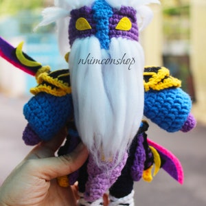May include: Crocheted plush toy of a character with a white beard, purple and blue clothing, and a yellow and black mask. The toy is holding a pink and yellow sword with a crescent moon design. The text "nhimconshops" is visible on the toy.