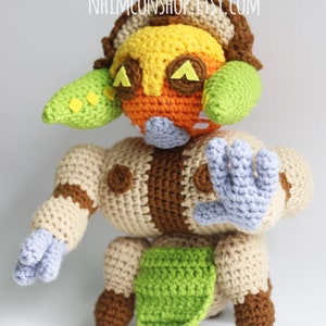 May include: Crocheted plush toy of a character with a brown, tan, orange, yellow, green, and purple color scheme. The character has a large head with a yellow and orange face, green ears, and brown eyes. The character has a tan body with brown accents and purple hands.