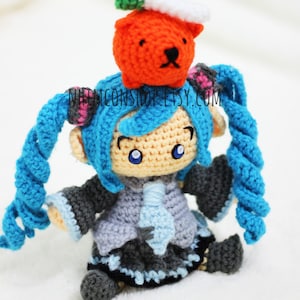 May include: A crocheted anime character with blue hair and a gray and blue outfit. The character is wearing a crocheted orange hat with a green leaf on top. The character is sitting on a white surface.