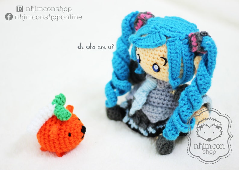 May include: A blue crocheted doll with long blue hair and a gray dress. The doll is holding a small orange crocheted pumpkin with green leaves. The doll is looking at the pumpkin and saying "eh who are u?"