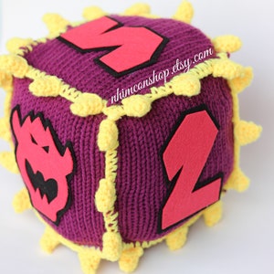 May include: A purple crocheted cube with yellow trim and red felt numbers. The cube has a  "rhimconshop.etsy.com" label on one side.