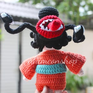 Puede incluir: Mu&ntilde;eca de ganchillo con una criatura monstruosa con boca roja, dientes blancos y lengua roja. La mu&ntilde;eca tiene brazos y pelo negros, y lleva un kimono naranja con una faja azul. El texto "honconshop" es visible.