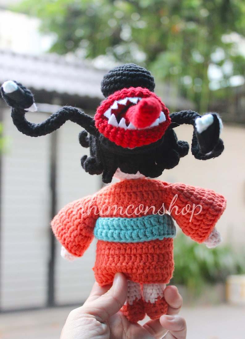 Puede incluir: Mu&ntilde;eca de ganchillo con una criatura monstruosa con boca roja, dientes blancos y lengua roja. La mu&ntilde;eca tiene brazos y pelo negros, y lleva un kimono naranja con una faja azul. El texto "honconshop" es visible.