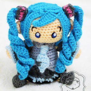 May include: A crocheted anime character with blue hair, a light blue and gray outfit, and large blue eyes. The character is standing on a white surface. The text "nhimconshop" and "nhimconshoponline" are visible in the image.