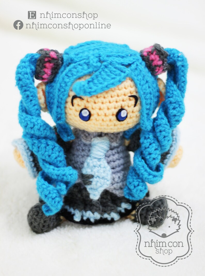 May include: A crocheted anime character with blue hair, a light blue and gray outfit, and large blue eyes. The character is standing on a white surface. The text "nhimconshop" and "nhimconshoponline" are visible in the image.