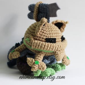 May include: A crocheted plush toy in the shape of a creature with a brown body, green accents, and a gray head with a black felt wing. The toy has black button eyes.
