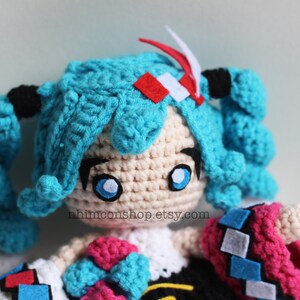 May include: Crocheted plush toy of a blue-haired anime character with large blue eyes. The toy is wearing a pink and blue outfit with a black and white checkered pattern. The toy is holding a blue bow in its hand. The text "nhmimconshop.etsy.com" is visible on the toy's chest.