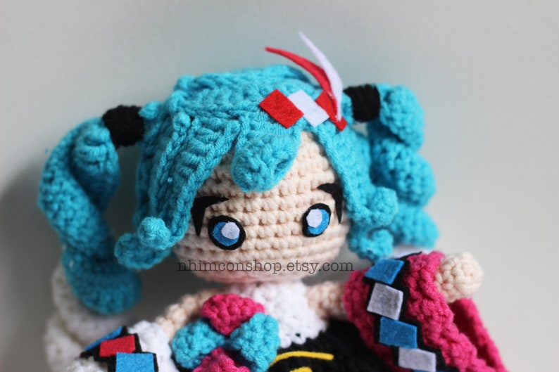 May include: Crocheted plush toy of a blue-haired anime character with large blue eyes. The toy is wearing a pink and blue outfit with a black and white checkered pattern. The toy is holding a blue bow in its hand. The text "nhmimconshop.etsy.com" is visible on the toy's chest.