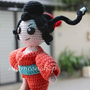 Puede incluir: Mu&ntilde;eca geisha de ganchillo, con un kimono rojo y una faja azul. La mu&ntilde;eca tiene pelo negro, cara blanca y ojos grandes y oscuros. El texto "nhimconshop" es visible en la ropa de la mu&ntilde;eca.