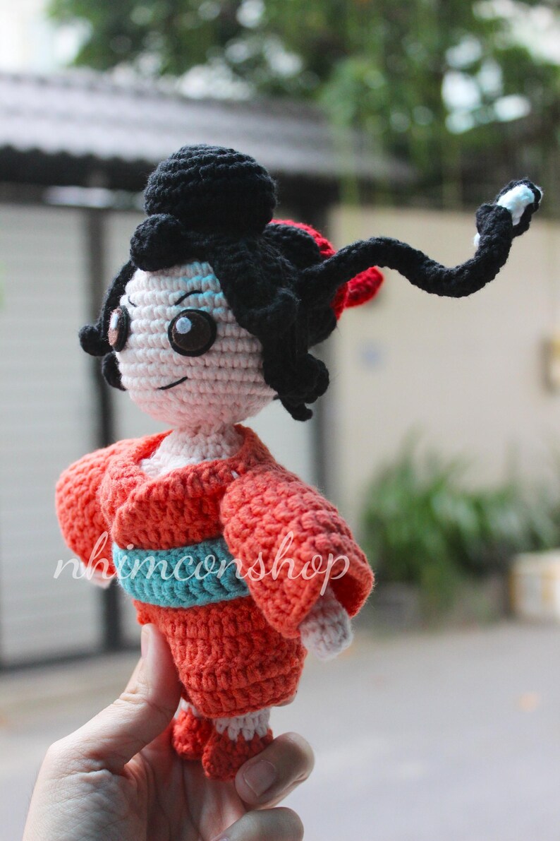 Puede incluir: Mu&ntilde;eca geisha de ganchillo, con un kimono rojo y una faja azul. La mu&ntilde;eca tiene pelo negro, cara blanca y ojos grandes y oscuros. El texto "nhimconshop" es visible en la ropa de la mu&ntilde;eca.