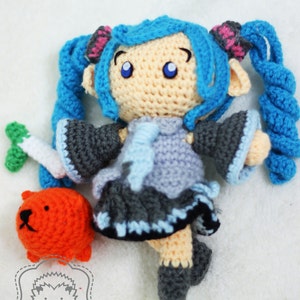 May include: A crocheted doll with blue hair in pigtails, wearing a gray and blue dress. The doll is holding a small orange crocheted animal. The doll is on a white background with the text "nhimconshop" and "nhimconshoponline" in black.