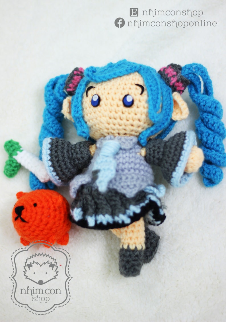 May include: A crocheted doll with blue hair in pigtails, wearing a gray and blue dress. The doll is holding a small orange crocheted animal. The doll is on a white background with the text "nhimconshop" and "nhimconshoponline" in black.