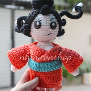 Puede incluir: Mu&ntilde;eca de ganchillo de estilo tradicional japon&eacute;s. La mu&ntilde;eca tiene cabello negro, ojos marrones y lleva un kimono rojo con una faja azul. El nombre de la mu&ntilde;eca, nhimconshop, est&aacute; escrito en el frente.