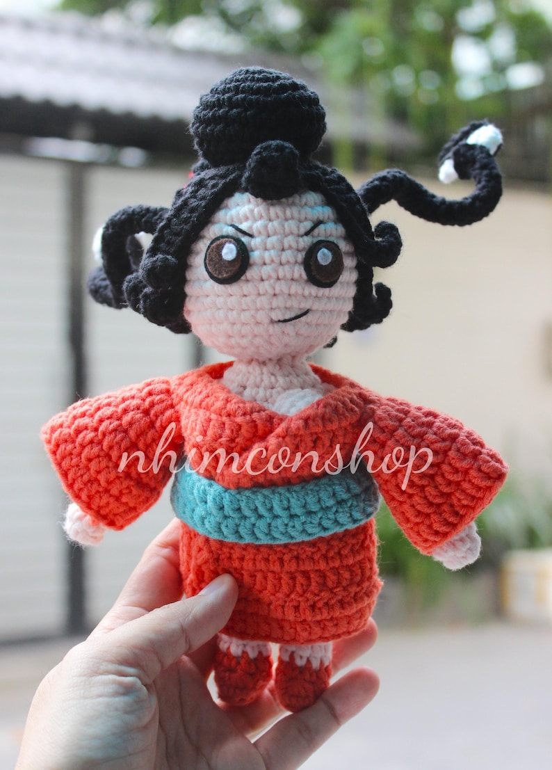 Puede incluir: Mu&ntilde;eca de ganchillo de estilo tradicional japon&eacute;s. La mu&ntilde;eca tiene cabello negro, ojos marrones y lleva un kimono rojo con una faja azul. El nombre de la mu&ntilde;eca, nhimconshop, est&aacute; escrito en el frente.