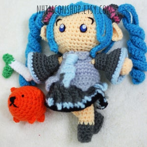 May include: Crocheted blue-haired doll with a grey and blue dress, holding a green carrot. The doll is standing on a white surface with a small orange crocheted animal beside it. The text "NHIMCONSHOP.ETSY.COM" is visible in the background.
