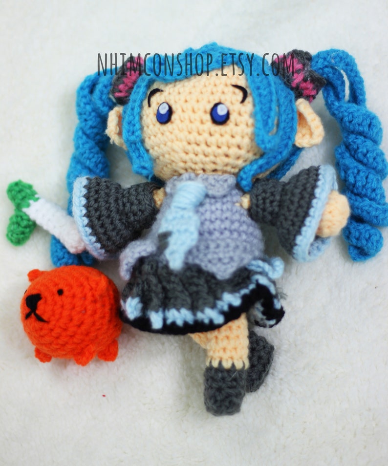 May include: Crocheted blue-haired doll with a grey and blue dress, holding a green carrot. The doll is standing on a white surface with a small orange crocheted animal beside it. The text "NHIMCONSHOP.ETSY.COM" is visible in the background.
