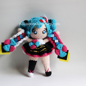 May include: Crocheted plush toy of a blue-haired anime character with pink and blue accents. The toy is wearing a black and yellow striped dress with a pink skirt. The toy has a yellow background with the text "nhimconshop.etsy.com".