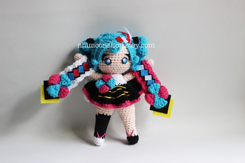 May include: Crocheted plush toy of a blue-haired anime character with pink and blue accents. The toy is wearing a black and yellow striped dress with a pink skirt. The toy has a yellow background with the text "nhimconshop.etsy.com".