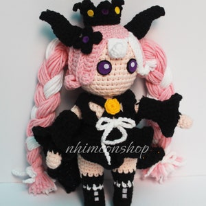 Puede incluir: Una muñeca de crochet con pelo rosa y blanco, un vestido negro y una corona negra. La muñeca tiene ojos morados y un botón amarillo en el pecho. La muñeca lleva zapatos negros con detalles blancos.